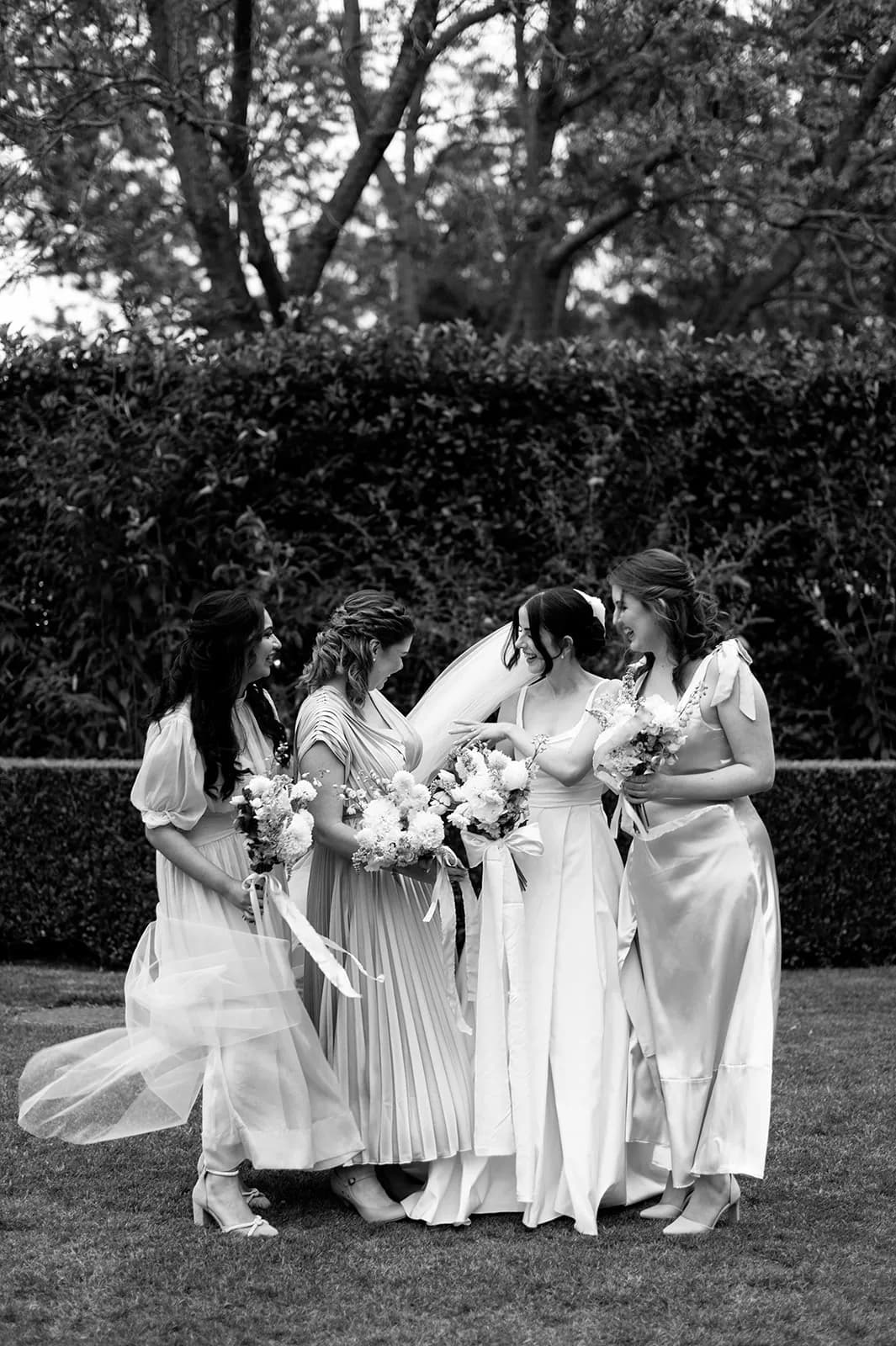 A black and white photo of four women standing in an outdoor garden, dressed in elegant gowns and holding bouquets. They are smiling and looking at each other, with trees and bushes in the background. The woman in the center is wearing a wedding dress with a veil.