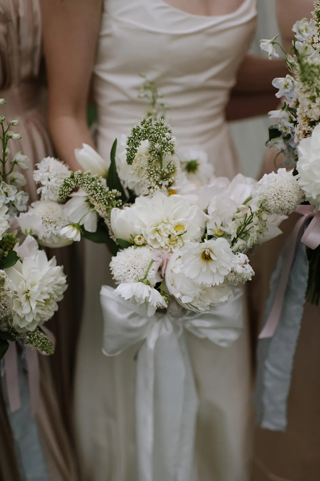 Three people in formal attire are holding floral bouquets. The central bouquet features white flowers with greenery, adorned with a white ribbon. The background is out of focus, highlighting the elegant and delicate arrangement of the flowers.