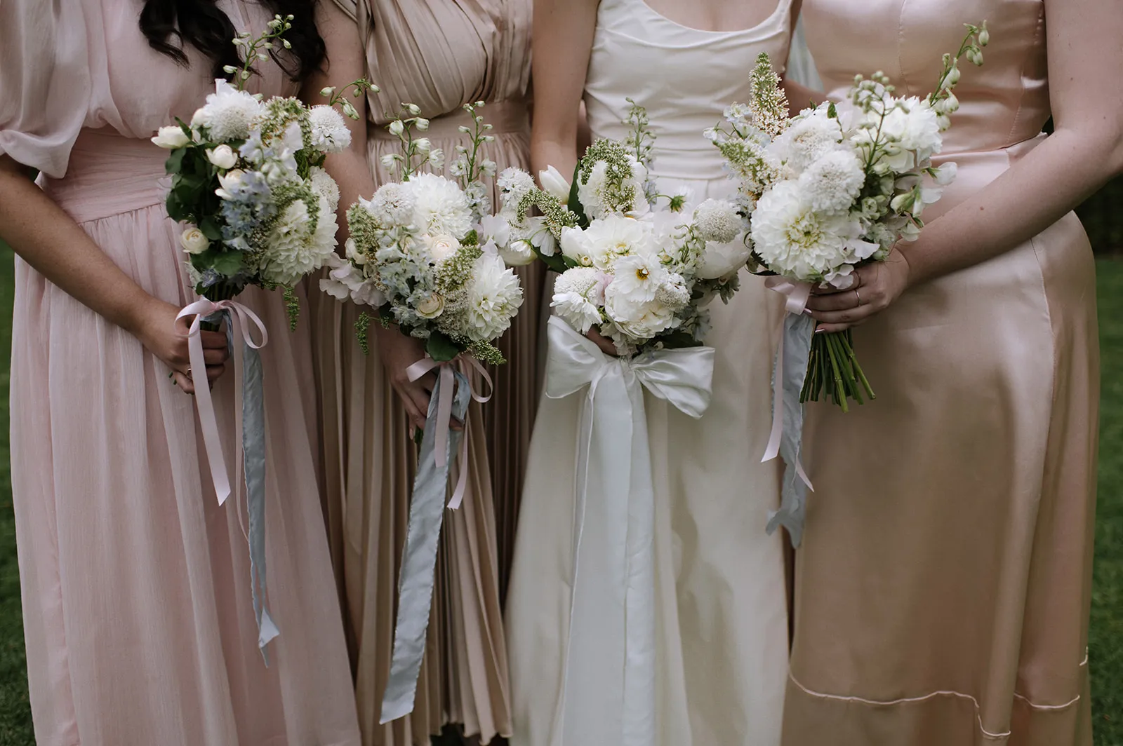 Three women in pastel-colored dresses hold bouquets of white flowers with greenery, tied with ribbons. Their faces are not visible, only their torsos and hands are shown. The background is a green, outdoor setting.