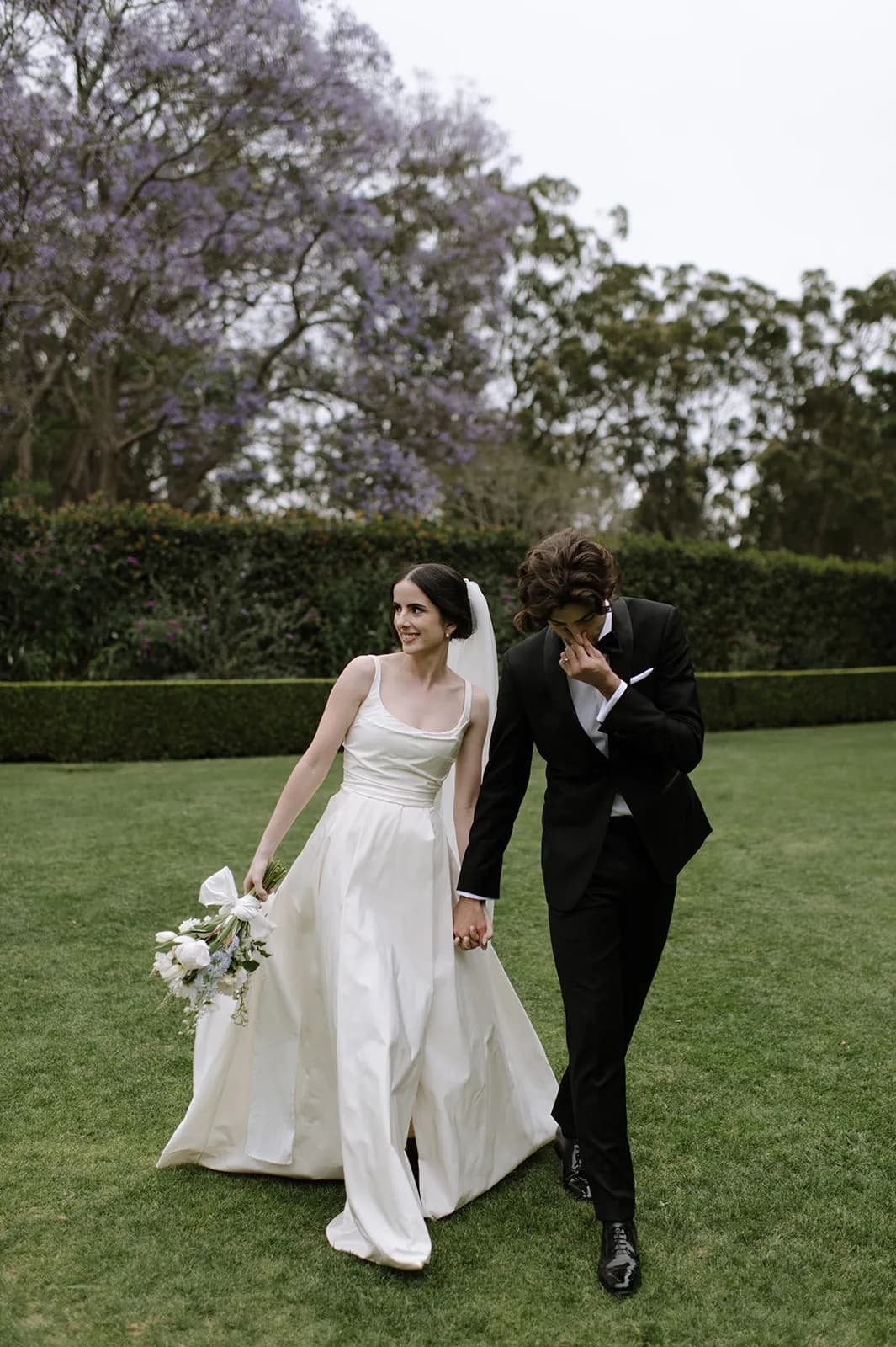 A bride and groom walk hand in hand on a grassy field. The bride is wearing a long white gown and holding a bouquet of flowers. The groom is dressed in a black suit and is covering his face with one hand, appearing emotional. Trees and bushes are in the background.