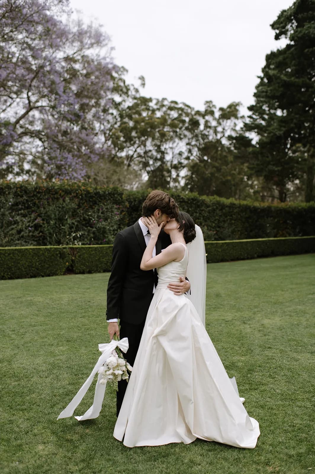 A newly married couple shares a kiss in a garden. The bride wears a white gown and veil, holding a bouquet of white flowers with ribbons, while the groom is dressed in a black suit, holding the bride's bouquet in his left hand. They stand on a well-manicured lawn.