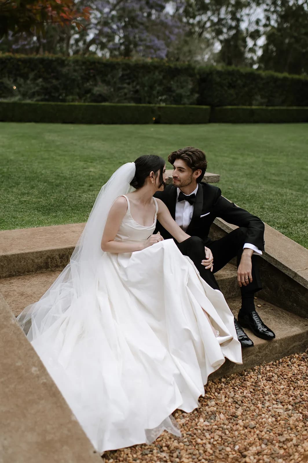 A bride in a white wedding dress and veil sits beside a groom in a black tuxedo, both sitting on the edge of a stone platform in a garden. They are gazing into each other's eyes, surrounded by lush greenery and a neatly manicured lawn.