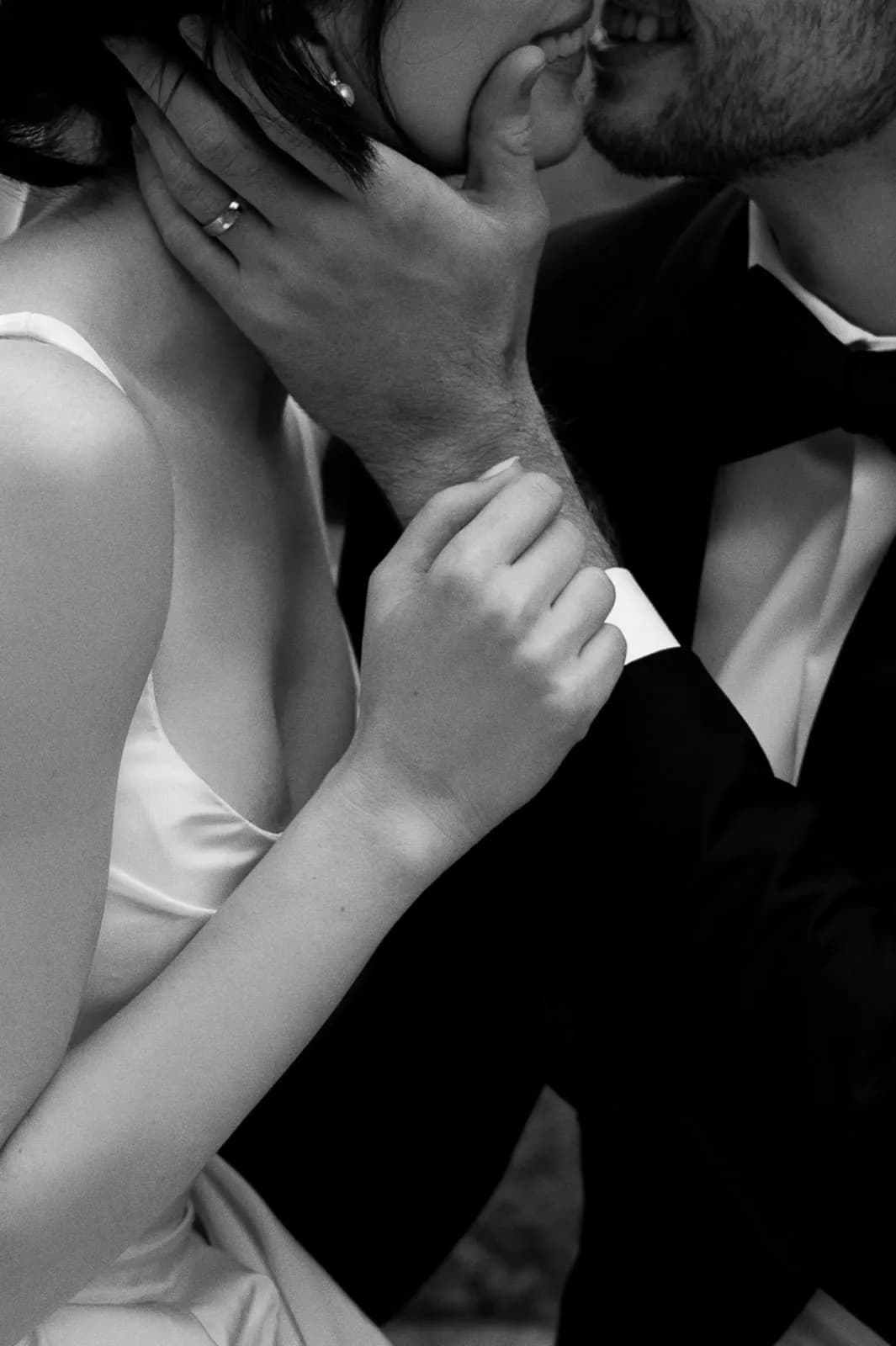 Black and white close-up of a couple in an intimate moment, with the person on the left wearing a sleeveless dress and the person on the right in a suit and bow tie. The person on the right gently holds the face of the other, who is wearing a ring. They appear to be about to kiss.