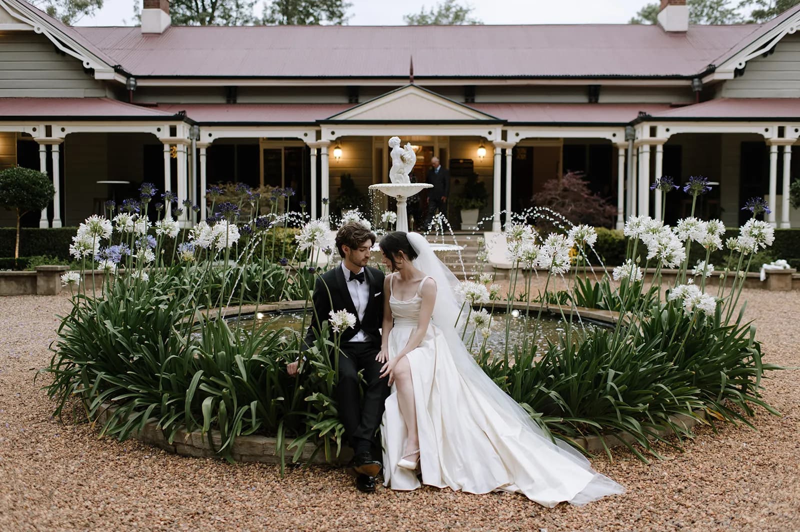 A bride and groom sit together by a circular fountain surrounded by plants and flowers. The bride wears a long white wedding dress and veil, while the groom is in a dark suit. They are in front of a large, elegant house with a red roof and white trim.