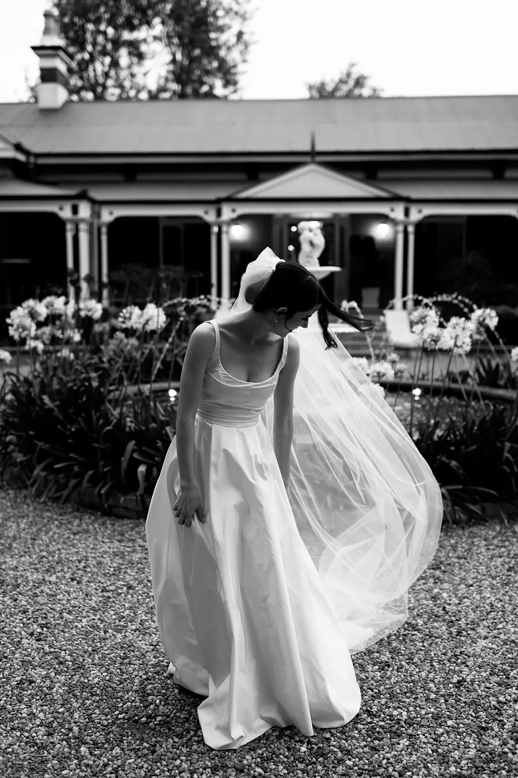 A bride in a flowing gown and veil stands on a gravel path in front of a building with large windows and a garden. She is slightly bent over and looking down. The black-and-white image highlights the elegance and serenity of the moment.