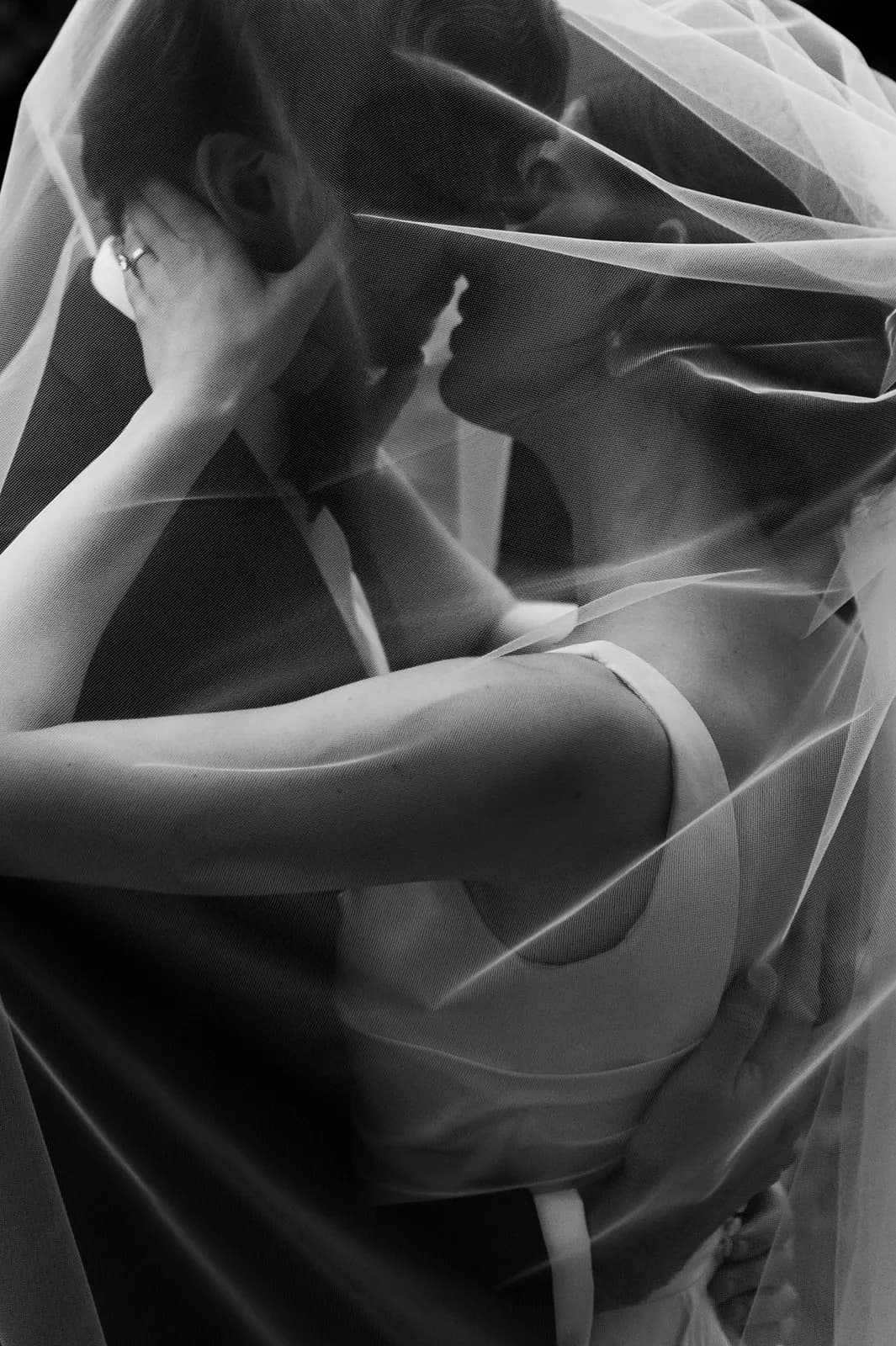 A black-and-white photo of a couple embracing and kissing under a sheer veil. They are close and intimate, with the bride's hand resting on the groom's neck and the groom's hand on the bride's waist. The bride wears a sleeveless dress and veil, and the groom is in a suit.