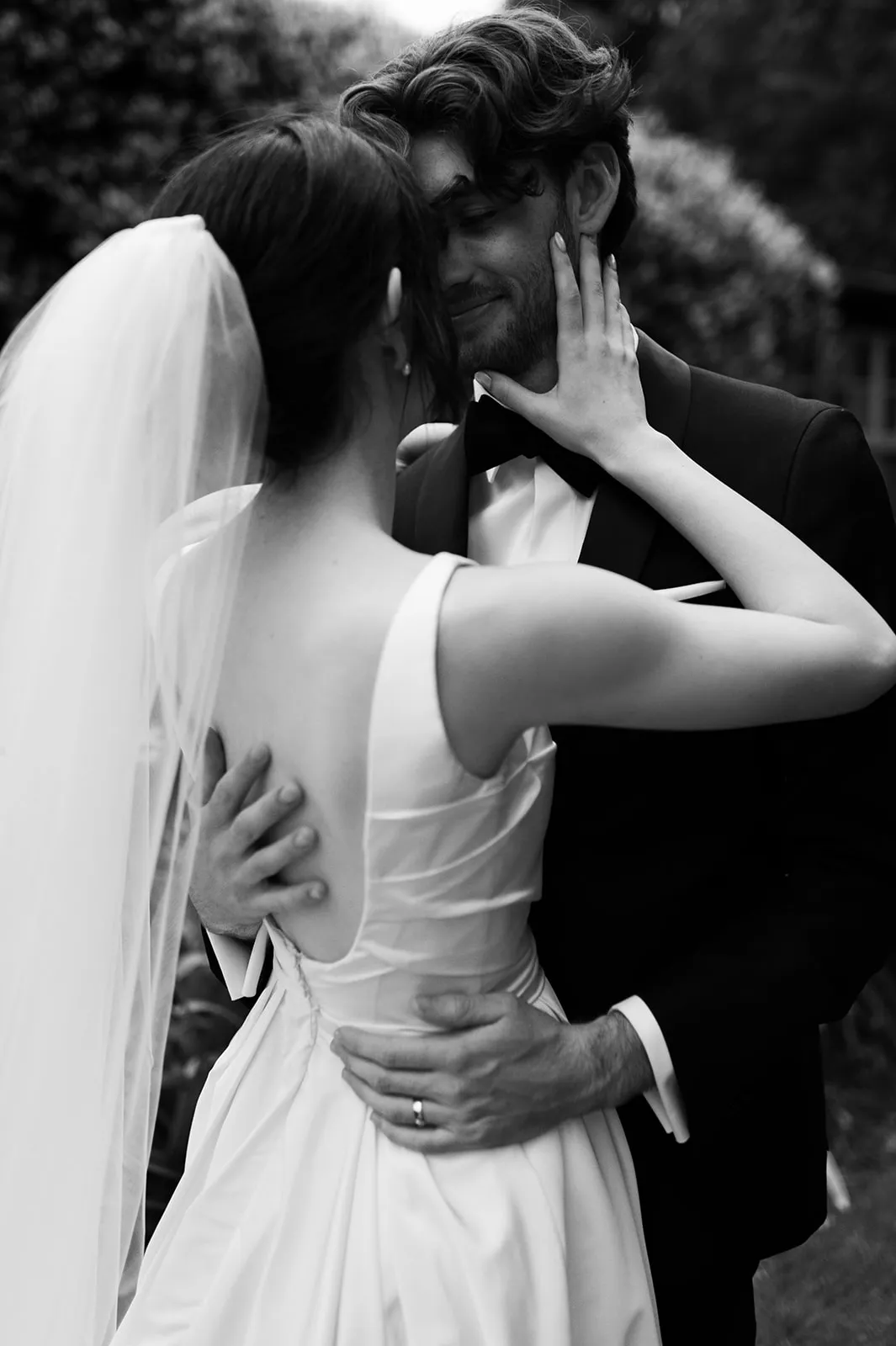 A black-and-white photograph captures a bride and groom sharing an intimate moment outdoors. The bride, wearing a veil and elegant dress, gently touches the groom's face while he holds her waist. They are both smiling and looking into each other's eyes.