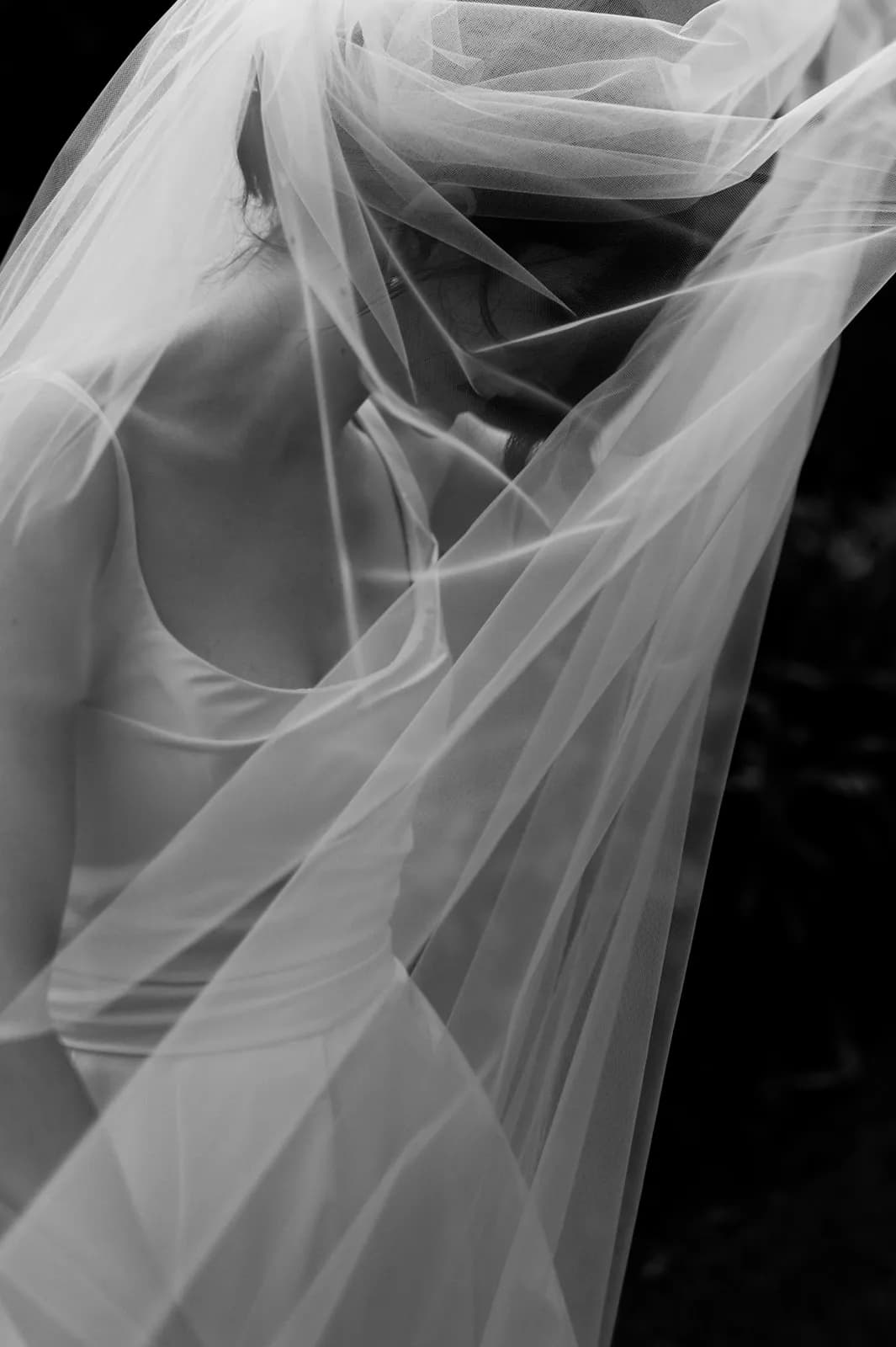 Black and white image of a bride wearing a flowing veil and sleeveless wedding dress. The veil covers her head and shoulders, and she looks down, creating an intimate and serene atmosphere.