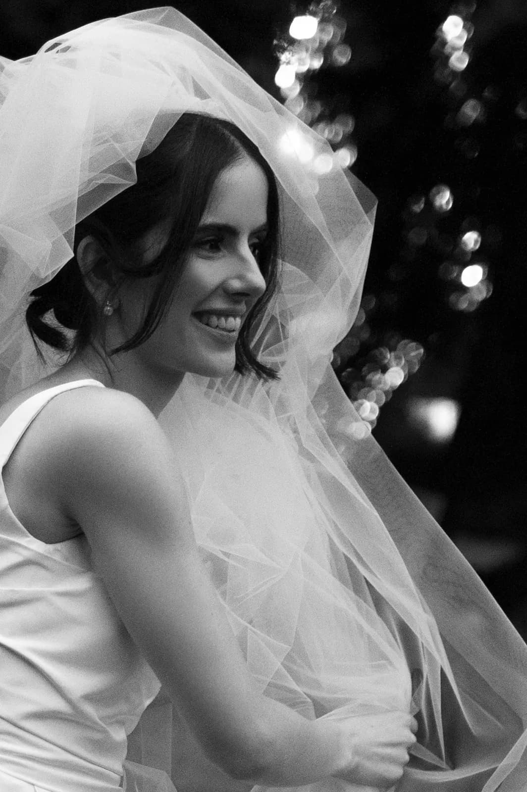 A black and white photo of a smiling bride with dark hair, wearing a veil and a sleeveless wedding dress. She appears to be outdoors, and the background features blurred lights or reflections.