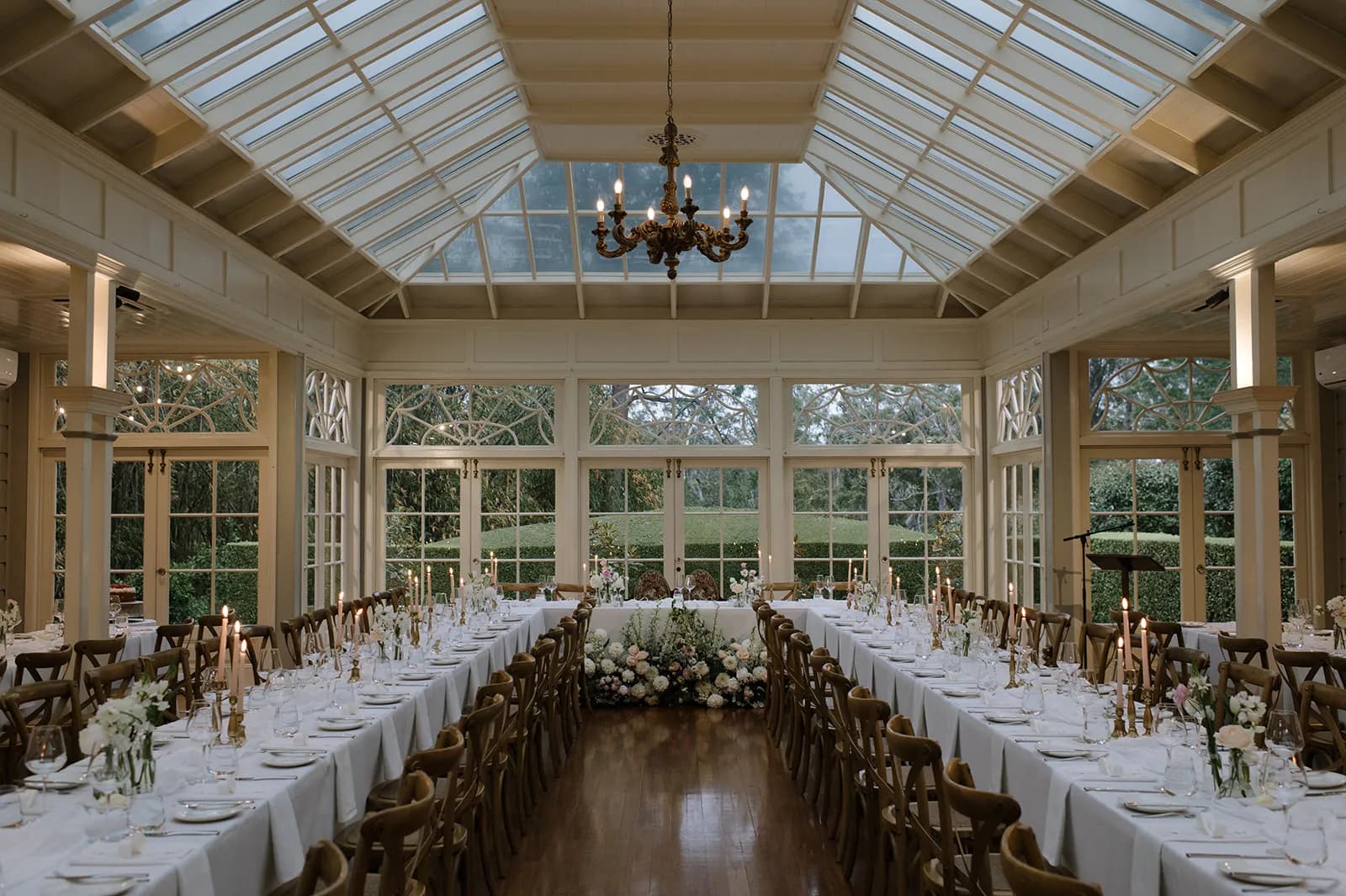 A beautifully decorated wedding reception venue with a glass ceiling and large windows overlooking a garden. Two long dining tables are adorned with white linens, elegant floral arrangements, and candles. A floral arrangement is centered where the couple will sit.