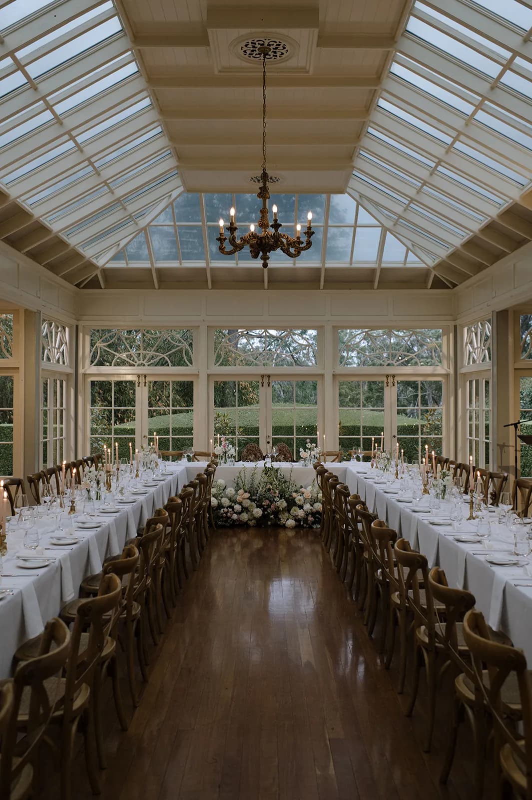 A beautifully set dining hall with long wooden tables adorned with white tablecloths and elegant table settings. The room features large, glass-paneled windows and a skylight ceiling. A vintage chandelier hangs from the ceiling, and there are floral arrangements in the center.