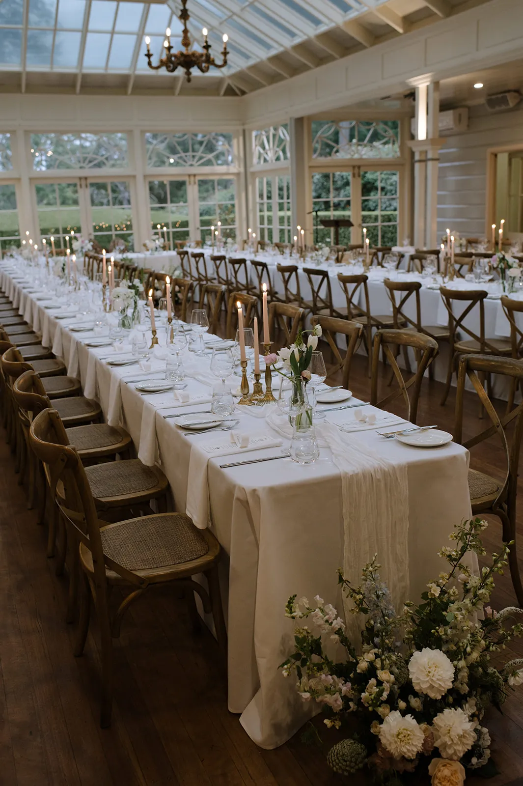 A beautifully set dining table in an elegant room with large windows and a glass ceiling. The long table features white tablecloths, candles, place settings, and floral centerpieces. Wooden chairs line both sides, creating a refined and festive atmosphere.