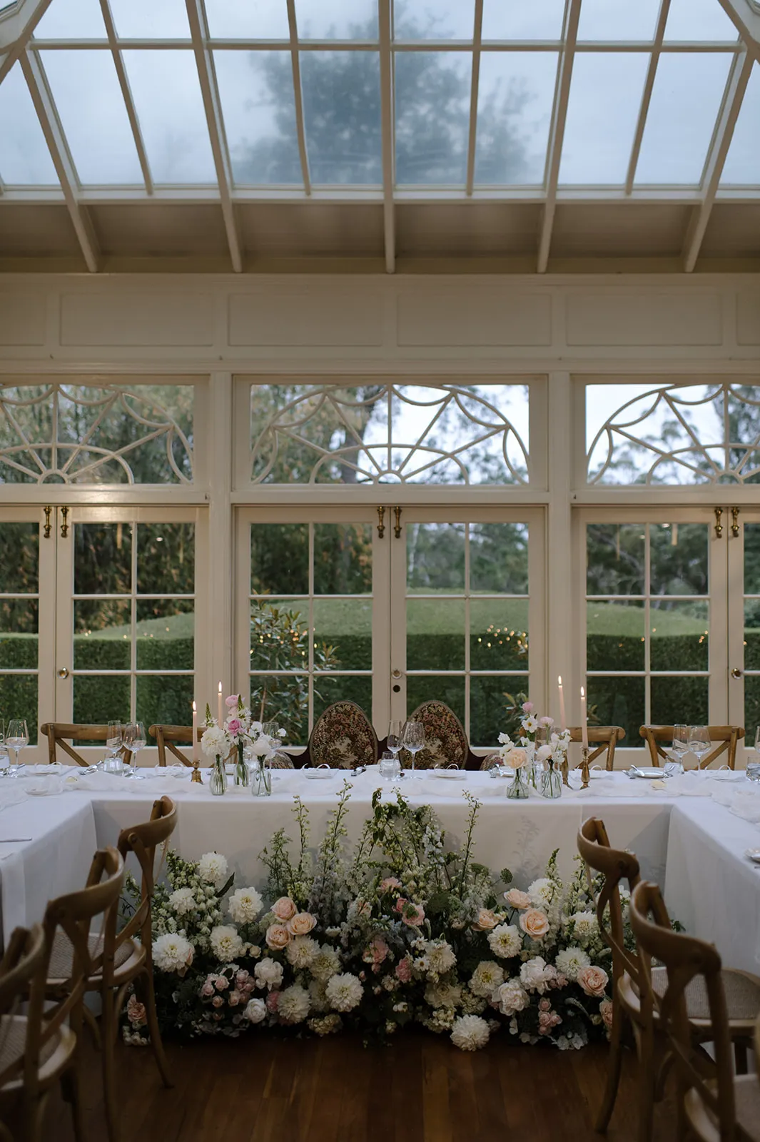 An elegant dining setup in a greenhouse with large windows and a glass ceiling. The table is adorned with white linens, floral arrangements, candles, and classic wooden chairs. The backdrop features a lush green garden visible through the windows.