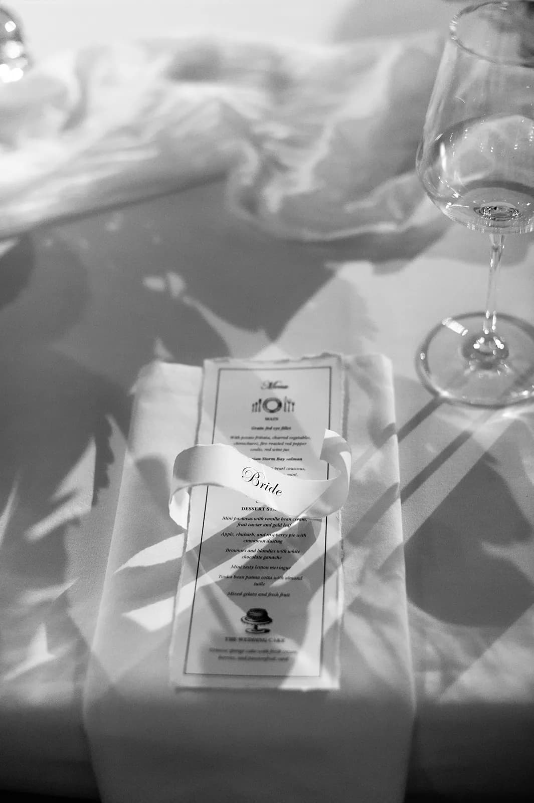 A black-and-white photo shows a neatly arranged place setting on a table. A napkin with a menu on top is held together by a ribbon labeled "Bride." An empty wine glass and soft shadows from floral decorations are on the table.