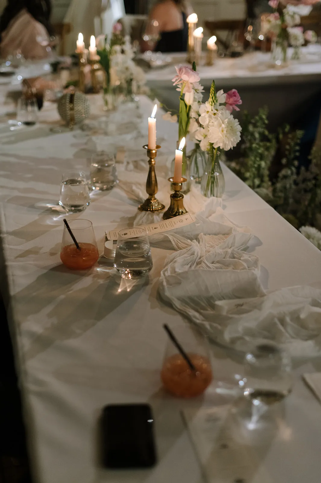 A dimly lit dinner table is set with white tablecloths, several lit candles, drinks with straws, clear glasses, and white flowers in vases. The atmosphere appears intimate and elegant. People are visible in the background, slightly out of focus.