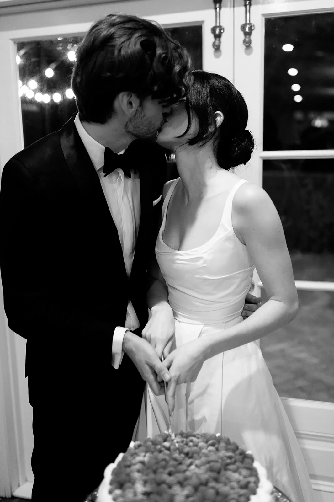 A newlywed couple is captured in a black-and-white photo, sharing a kiss while cutting their wedding cake together. The groom, dressed in a tuxedo, and the bride, in a sleeveless wedding gown, hold the cake knife together.