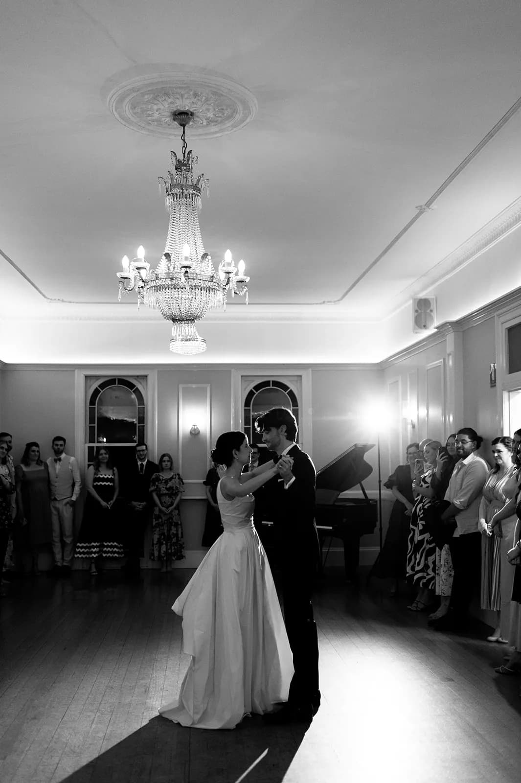 A bride and groom share their first dance in a elegantly decorated room, illuminated by a large chandelier. Guests stand around the room watching the couple. The image captures a timeless and intimate moment of the wedding celebration.