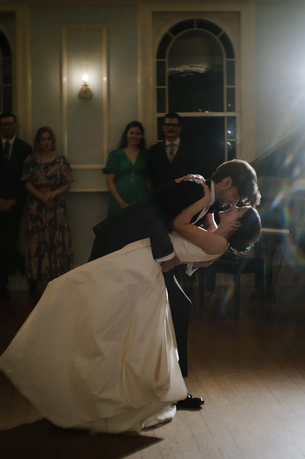 A bride and groom share a dance at their wedding, with the groom dipping the bride under a spotlight. They are surrounded by guests who are watching and smiling. The bride wears a white wedding dress, and the groom is in a dark suit. The room has elegant decor.