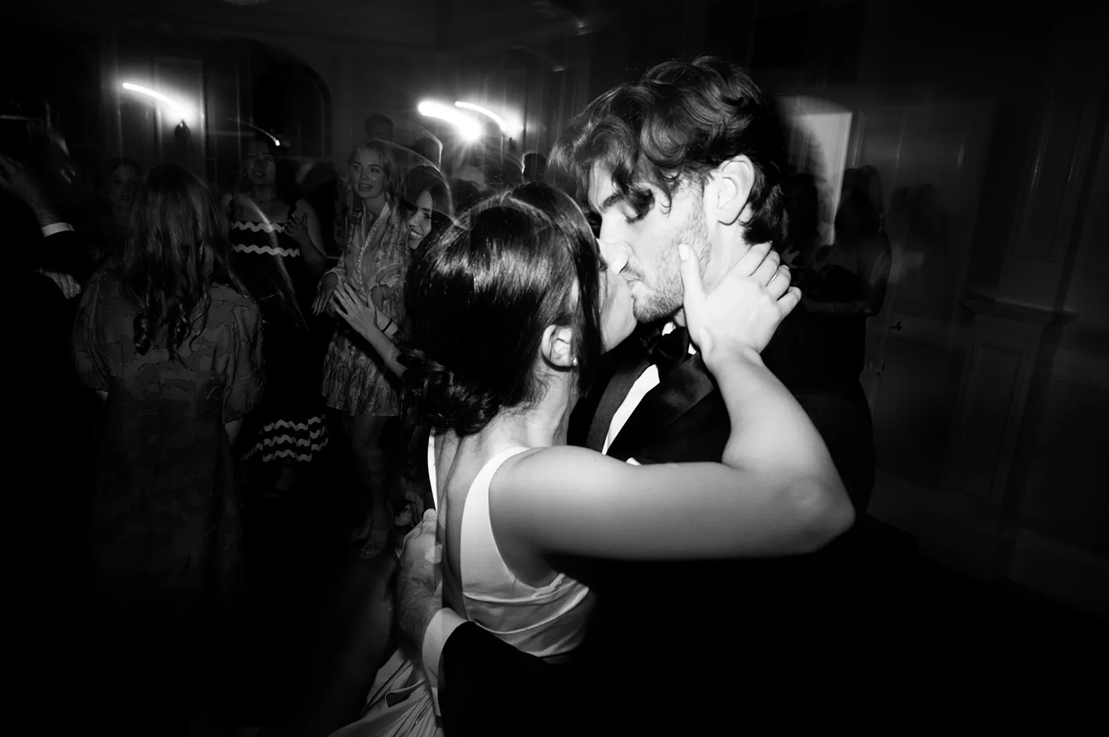 A black-and-white photograph shows a couple sharing an intimate dance, with the woman’s hands around the man's neck and their faces close together. They are surrounded by other people who are also dancing and socializing in the background.