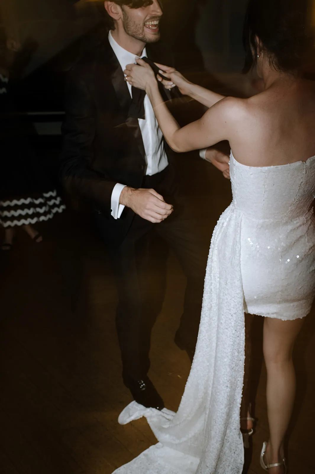 A couple dances at a formal event. The man is wearing a black suit and white shirt, and the woman is in a strapless white dress with a long train. They are smiling and enjoying the moment on the dance floor, with a blurred background suggesting movement.