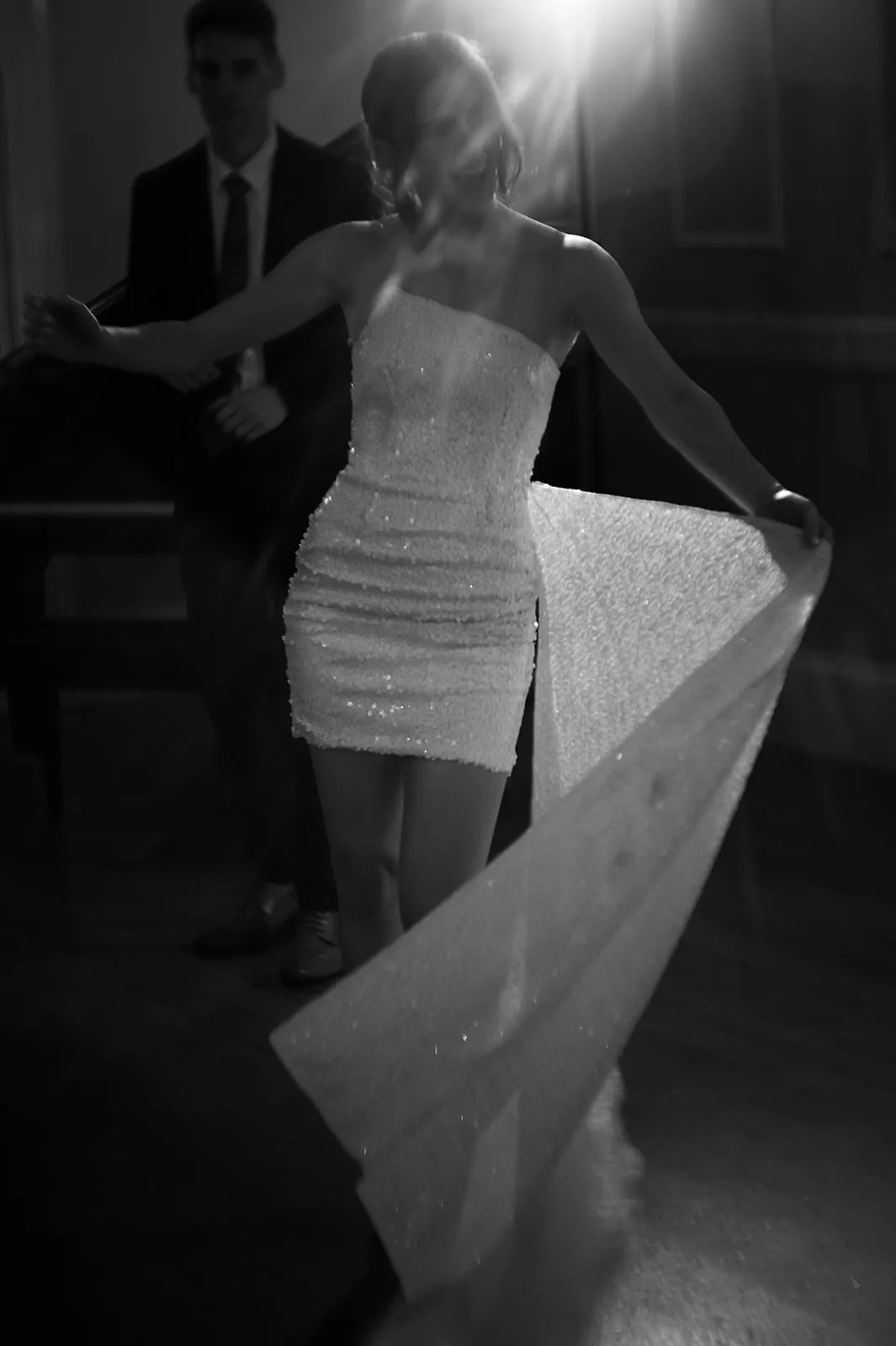 A black and white photo of a person wearing a strapless, glittery dress walking with a long, sparkling fabric trailing behind. Another person dressed in a suit is standing in the background, slightly out of focus. A bright light shines from the upper right.