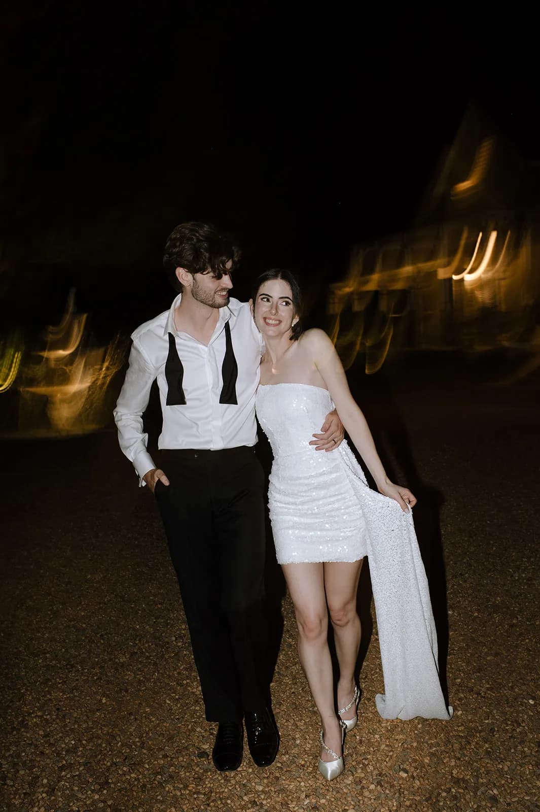 A couple walks together at night. The man, in a semi-formal outfit with an untied bow tie, has his arm around the woman who is wearing a short white dress and heels. The background is blurred, highlighting the couple's warm expressions.