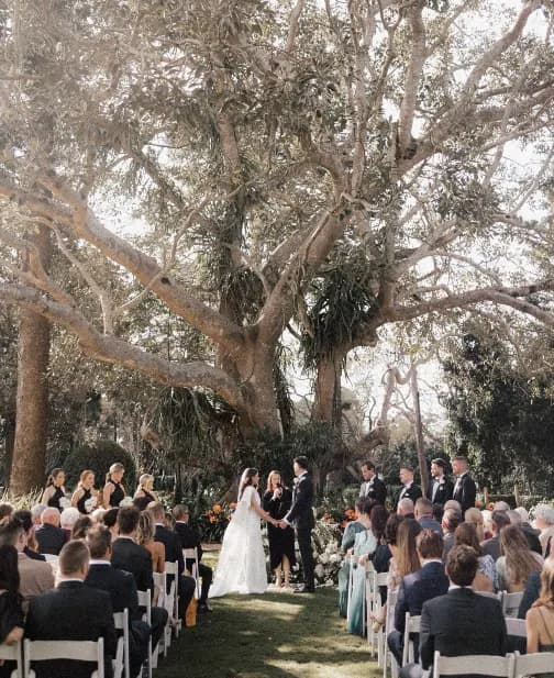 A couple stands under a large tree during an outdoor wedding ceremony. They are surrounded by their bridal party, who are dressed in formal attire. Guests are seated on white chairs in rows, observing the ceremony. It is a sunny day with natural light filtering through the tree leaves.