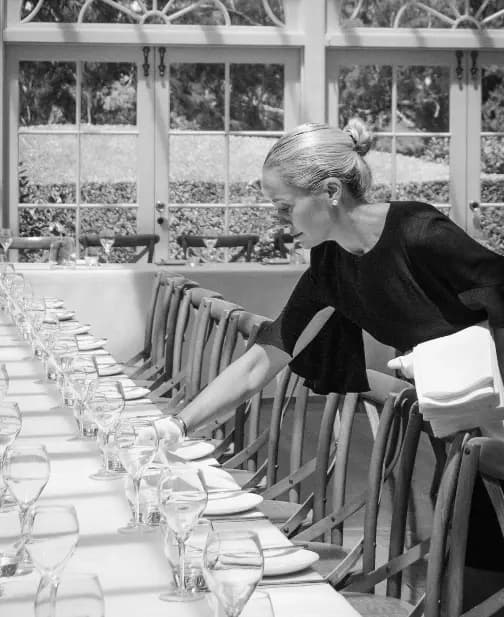 A person with their hair in a bun is arranging plates and wine glasses on a long, elegantly set dining table. The table has a white tablecloth and wooden chairs. Sunlight is streaming in through the large windows in the background.