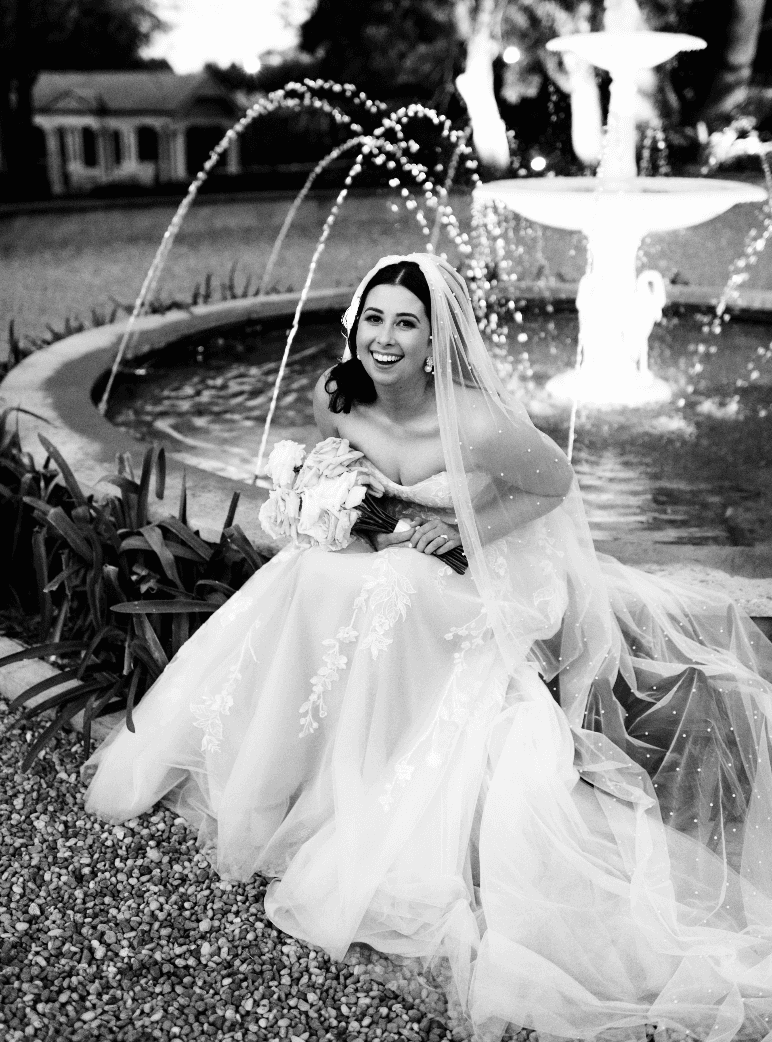 A bride in a white wedding dress and veil sits smiling in front of a fountain with water arcs. She holds a bouquet of flowers and looks joyful. The scene is captured in black and white, with a garden setting in the background.