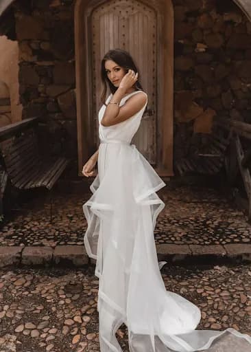 A woman in a flowing white dress stands outdoors against a rustic wooden door and stone wall backdrop. She has long, dark hair and touches her face with one hand, projecting elegance and grace. Benches line the sides, and the ground is cobblestone.