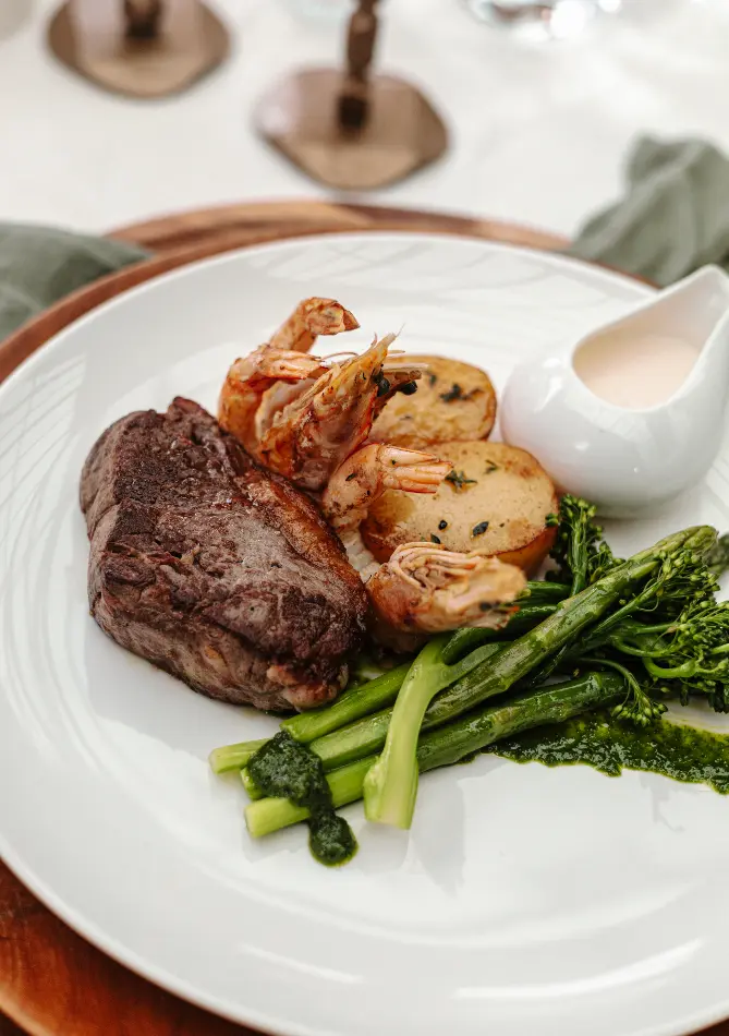 A plate of surf and turf featuring a juicy steak, two grilled shrimp atop roasted potatoes, and a side of broccolini. A small white pitcher containing a creamy sauce is on the plate as well. The background shows a blurred table setting.