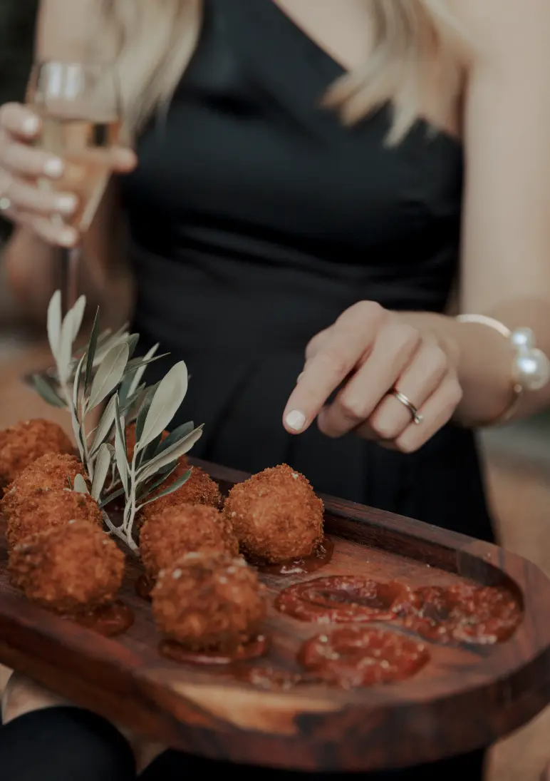 A person dressed in a black outfit holds a wooden tray with several golden-brown, fried appetizers garnished with some greens. They are dipping one of the appetizers into a red sauce. In their other hand, they hold a glass of champagne.