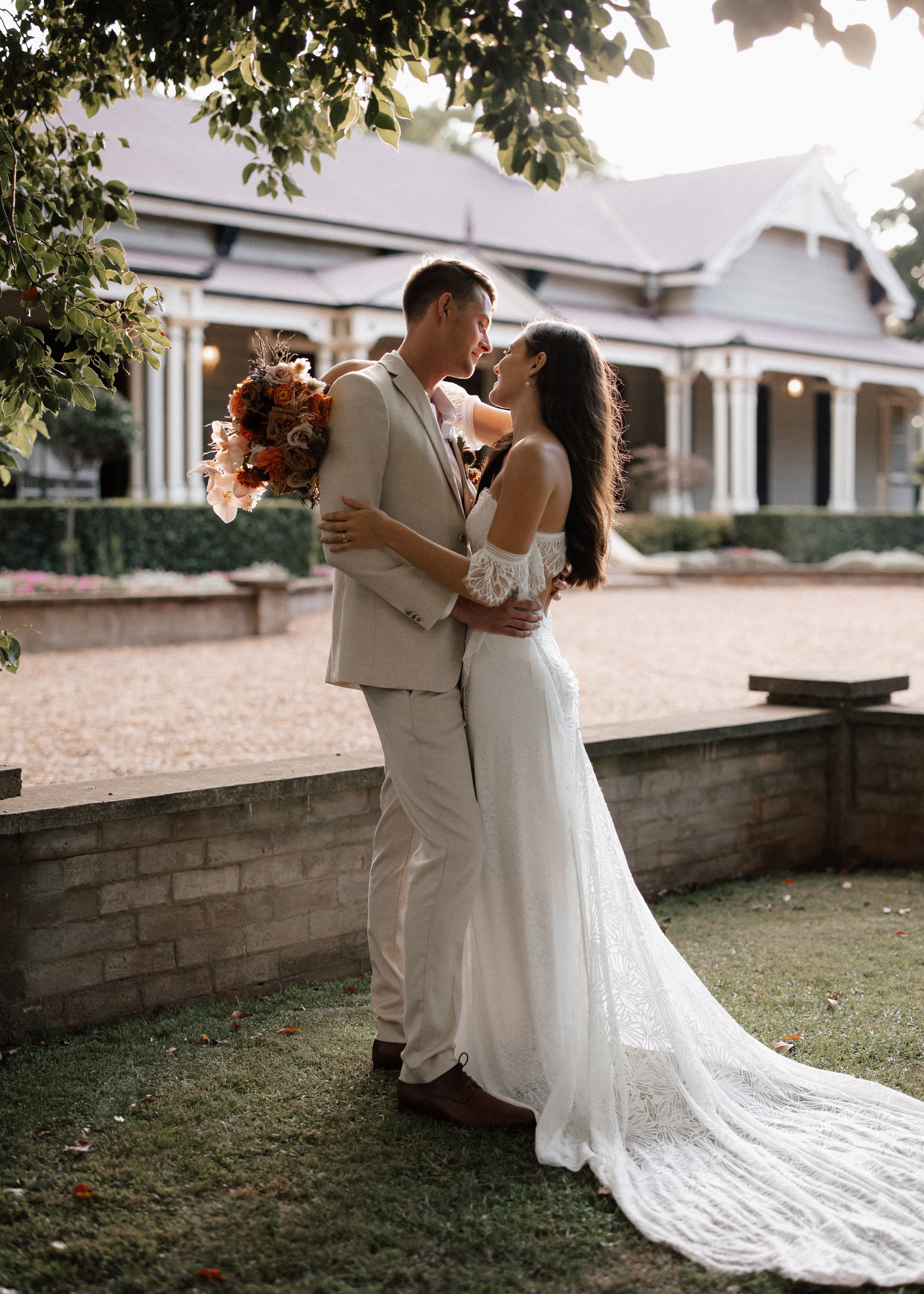 A bride and groom joyfully embrace in front of a vintage house. The groom holds a bouquet of flowers and wears a light beige suit, while the bride is in a long, white lace wedding dress. Sunlight filters through the trees, casting a warm, romantic glow on the scene.