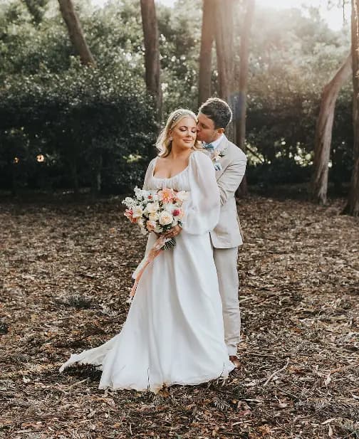 A bride in a white gown holding a bouquet of flowers stands in front of a groom in a light-colored suit. They are in a wooded area with sunlight filtering through the trees. The groom embraces the bride from behind as they both smile.