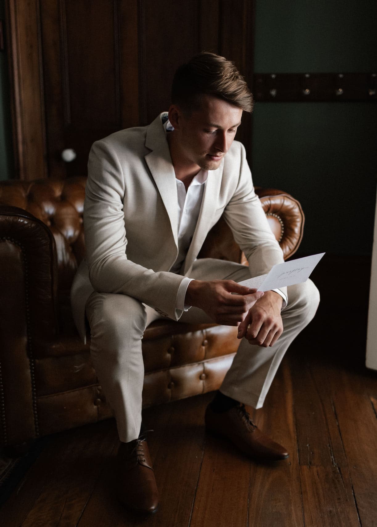 A man in a light beige suit sits on a brown leather couch, reading a piece of paper. He has short brown hair and is leaning slightly forward, absorbed in the contents of the paper. The room has dark wooden floors and a dark green wall.