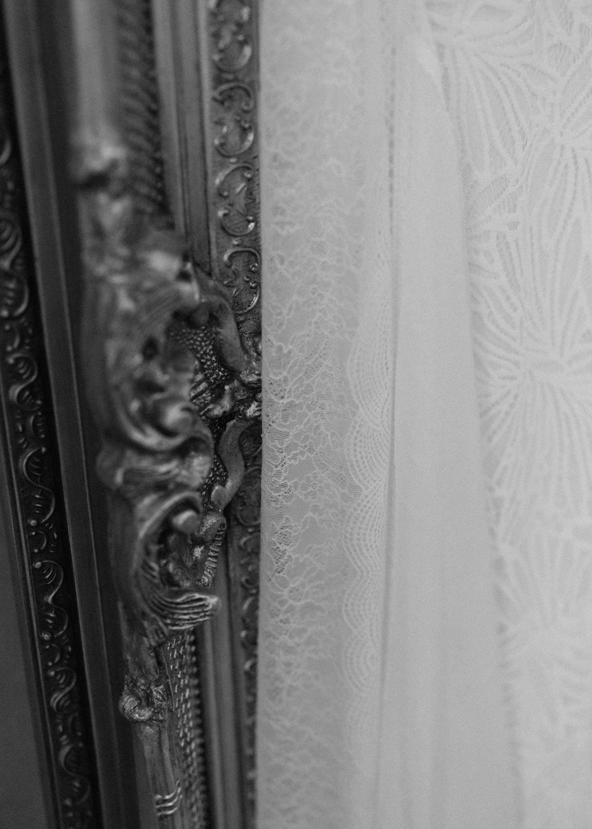 Black and white close-up of an ornate vintage frame next to a delicate lace fabric. The frame features intricate carvings, and the lace shows a subtle floral pattern, emphasizing the textures and details of both materials.