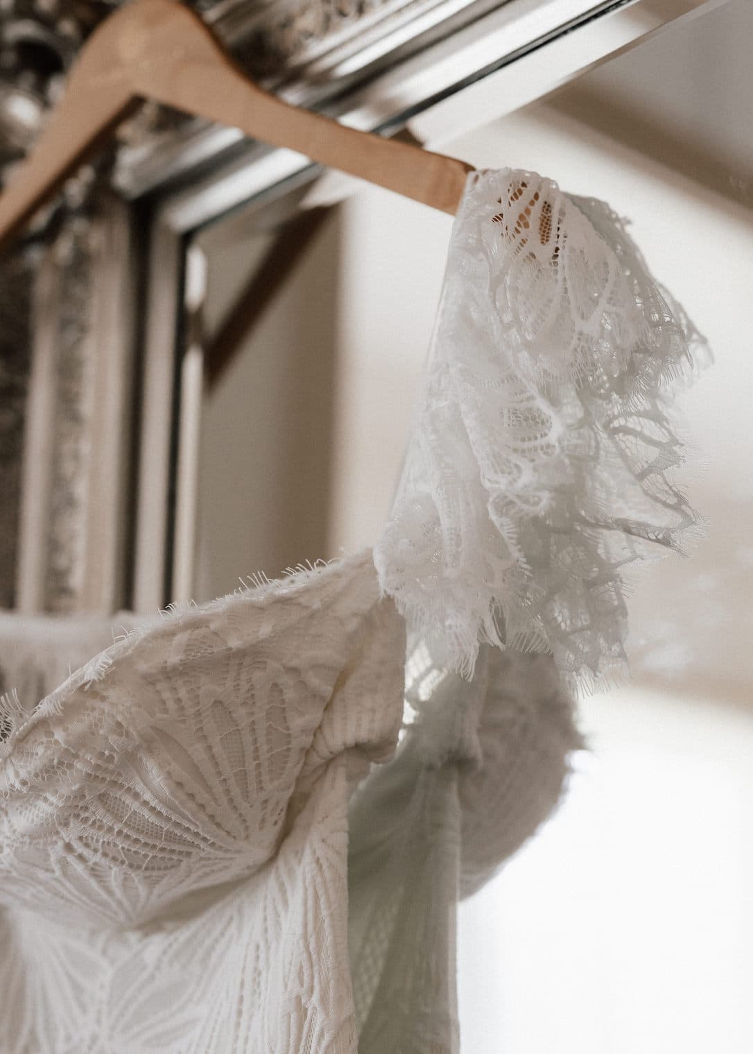 Close-up of a white lace wedding dress hanging on a wooden hanger in front of a mirror. The intricate lace detail and delicate fabric are emphasized, showcasing the craftsmanship of the garment. The soft lighting adds a romantic ambiance to the scene.