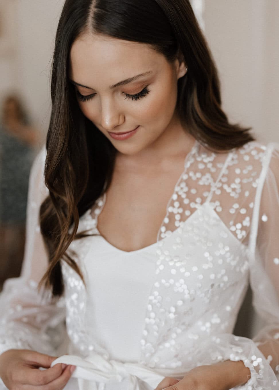 A woman with long brown hair is looking down and smiling slightly. She is wearing a white outfit with a sheer, embroidered overlay that has floral patterns. She appears to be tying a ribbon at her waist. The background is softly blurred, highlighting her features.