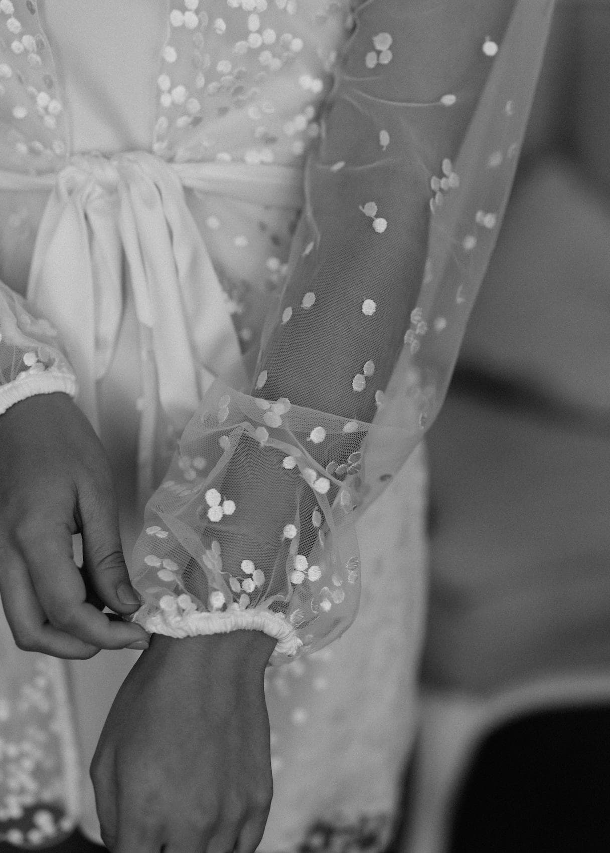 Black and white close-up of a person adjusting the sleeve of a sheer, sequined garment tied with a satin ribbon at the waist. The sleeves have elastic cuffs, and the intricate detail of the fabric is highlighted.