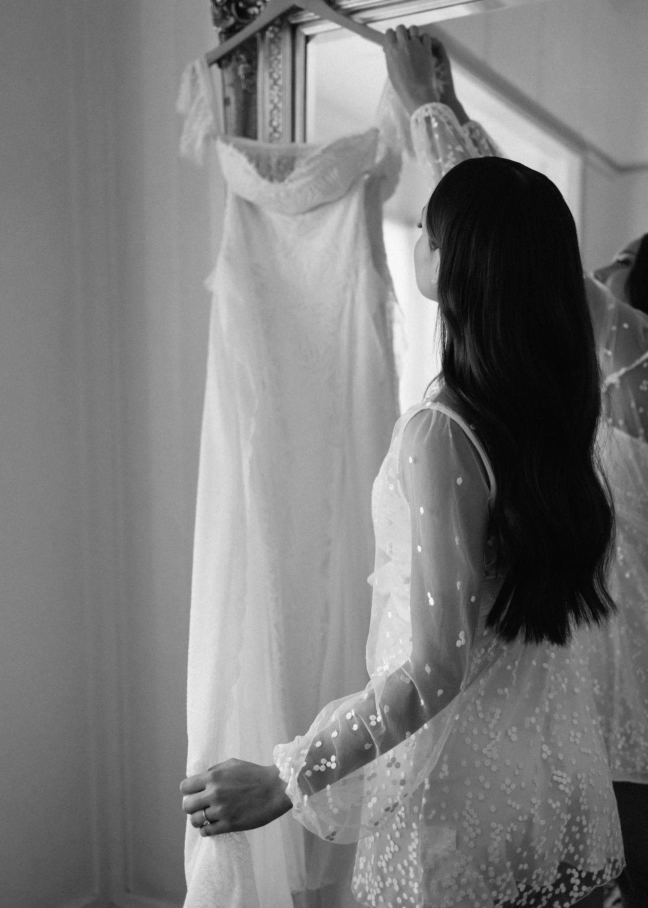 A woman with long, dark hair stands in front of a mirror, holding a white lace wedding dress that hangs on a rack. She is wearing a sheer, polka-dotted blouse and has her back to the camera. The scene is captured in black and white.