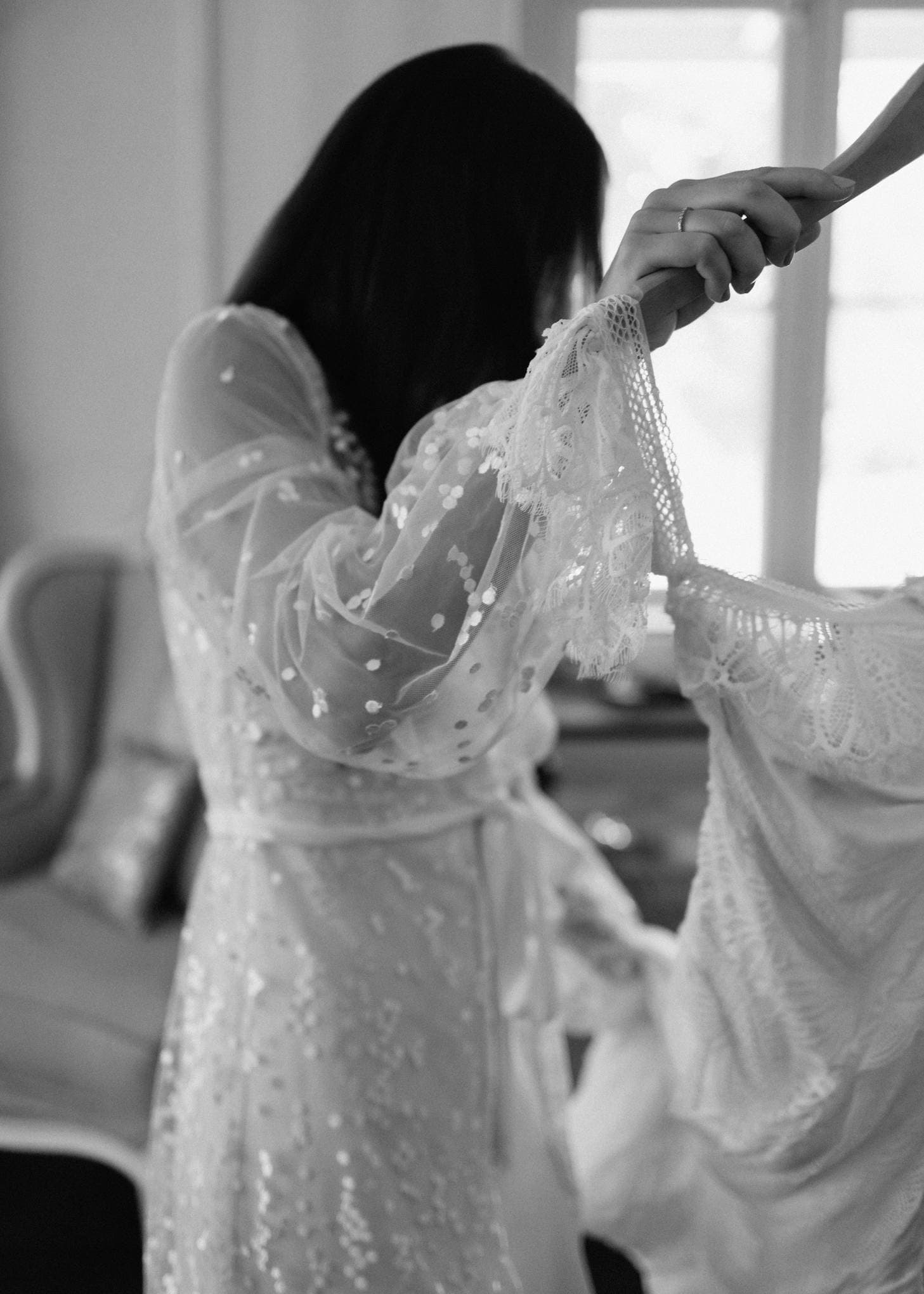 A woman with long dark hair, seen from the side, wearing a sheer, long-sleeved garment with lace and polka dots. She holds a white lacy dress on a hanger. The background shows a cushioned chair and a window. The image is in black and white.