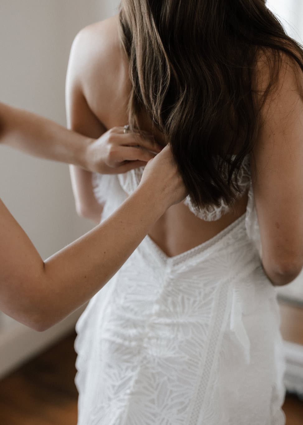 A woman in a white lace wedding dress is seen from the back, her long brown hair cascading down. Another person is helping to fasten the dress. The room has soft lighting and wooden flooring, adding to the intimate and tender moment.