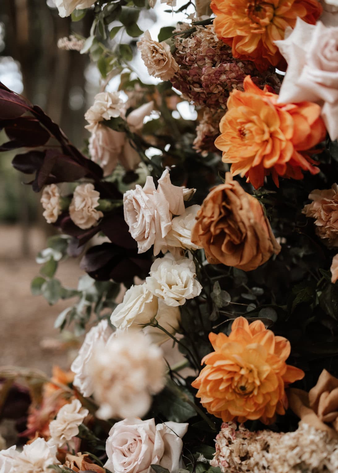 A close-up of an outdoor floral arrangement featuring a mix of orange dahlias, white roses, peach-colored roses, and other assorted blooms. The background includes blurred greenery and trees.