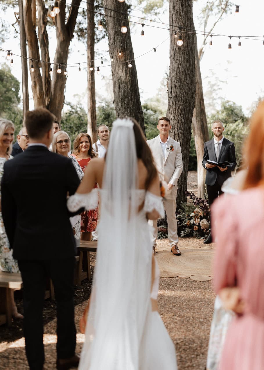 A bride in a long white dress and veil walks down the aisle, accompanied by a man in a dark suit. They are surrounded by guests standing in a wooded outdoor setting. The groom, dressed in a light suit, waits at the altar, along with an officiant holding a book.