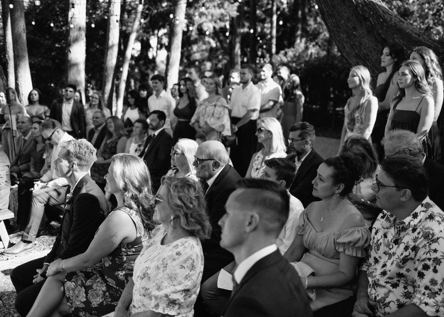 A group of people, dressed in formal and semi-formal attire, sit and stand closely together outdoors in a wooded area. The sun shines through the trees, casting dappled light on them. Some people are smiling, while others appear engaged in what seems to be an event or ceremony.