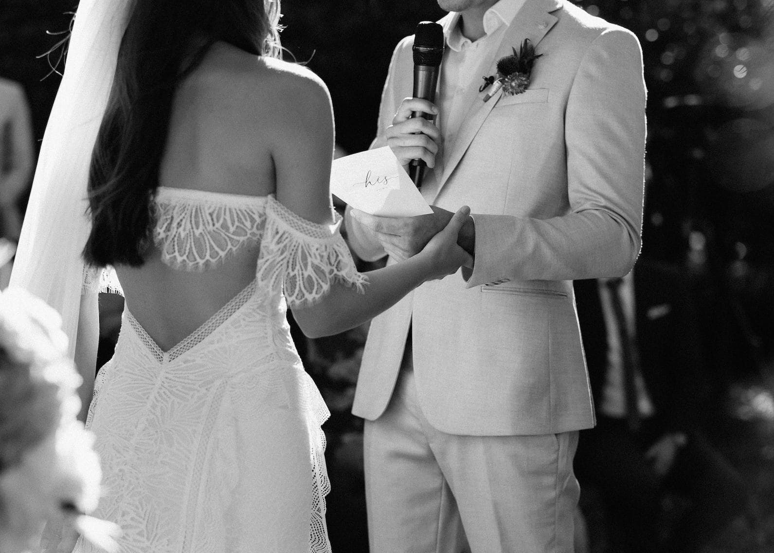 A black-and-white photo captures a wedding ceremony where the groom, dressed in a light suit, holds a microphone and reads vows from a card while holding the bride’s hand. The bride, in a lace dress with an off-shoulder design, stands facing him.