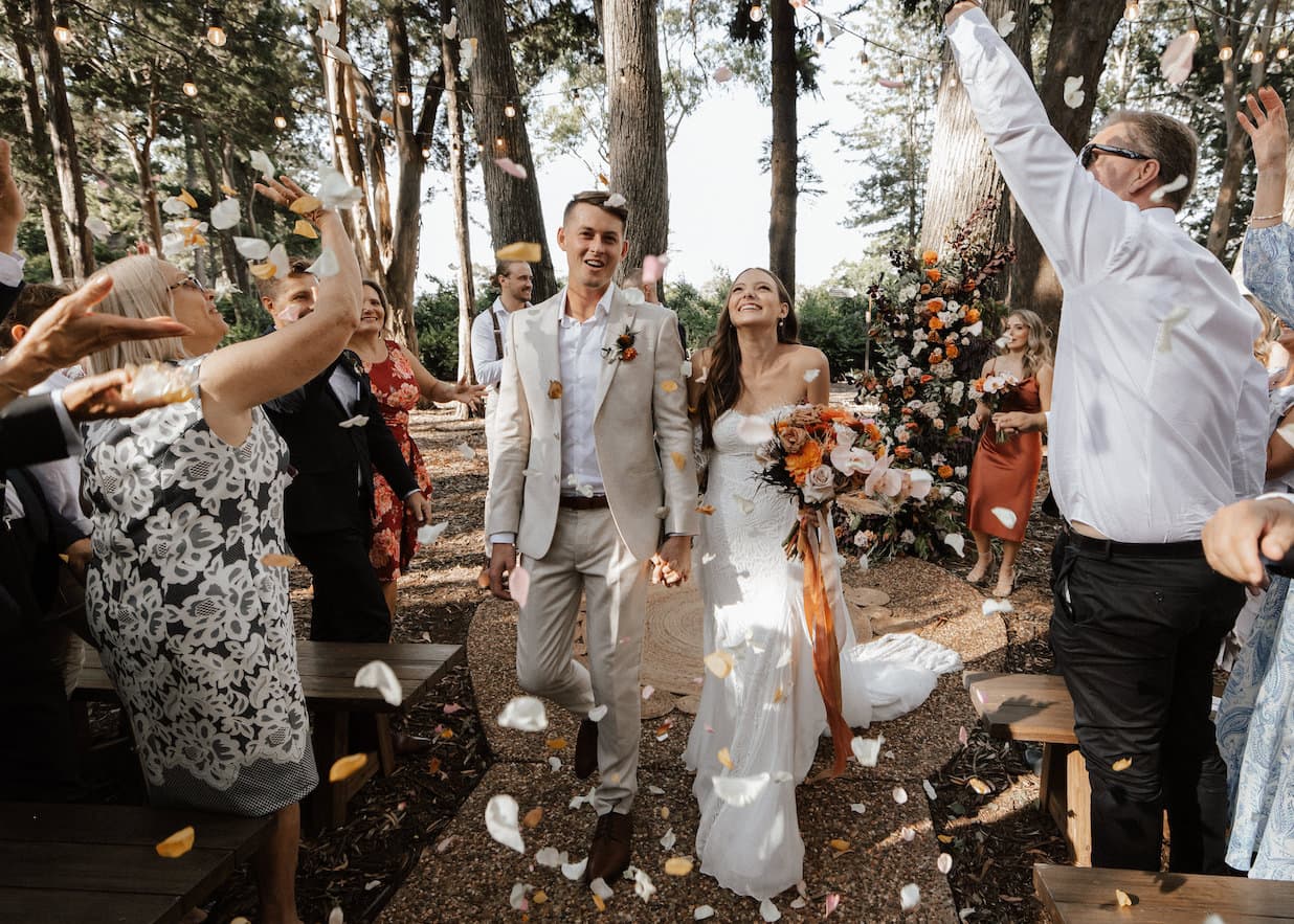 A happy couple in wedding attire walks down an outdoor aisle as guests throw flower petals. The bride holds a bouquet of flowers and wears a white wedding dress, and the groom is dressed in a light-colored suit. Trees and greenery fill the background.