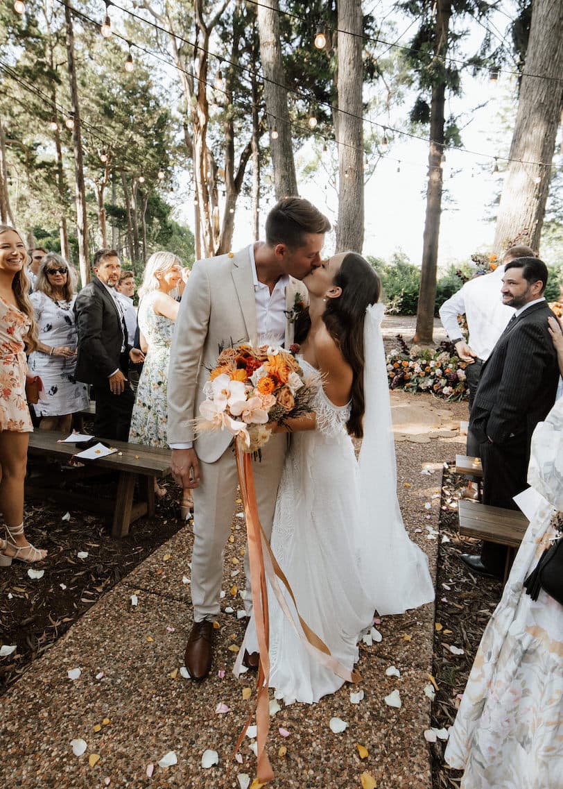 A newlywed couple shares a kiss outdoors surrounded by guests. The groom, in a light suit, and the bride, in a white lace gown and veil, hold a bouquet of orange and white flowers with long ribbons. Guests stand and smile among trees adorned with string lights.