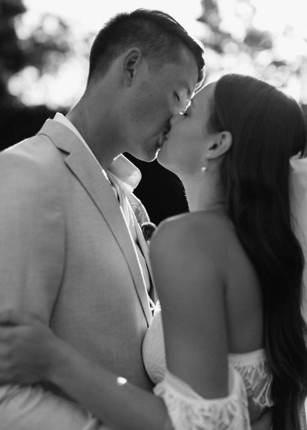 A black and white photo of a couple sharing a kiss. The man is wearing a light-colored suit while the woman is in a white off-shoulder dress. They are outdoors with blurred trees and sunlight in the background.