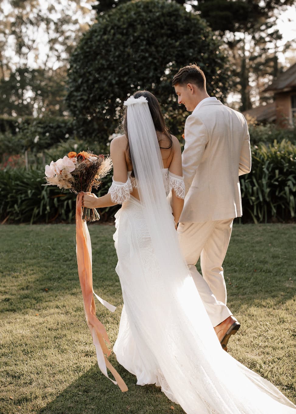 A bride and groom walk hand in hand through a lush garden. The bride wears a flowing white dress with a long veil, holding a bouquet of flowers with long ribbons. The groom is dressed in a beige suit. Sunlight filters through the trees, creating a warm, serene atmosphere.